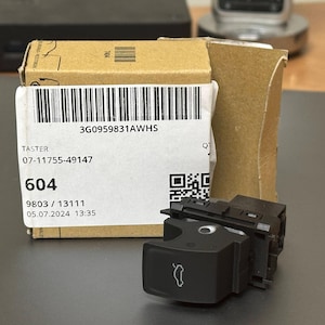 Könnte beinhalten: Ein brauner Karton mit einem weißen Etikett. Das Etikett enthält einen Barcode, den Text "3G0959831AWHS", "TASTER", "07-11755-49147", "604", "9803 / 13111" und "05.07.2024 13:35". Vor der Box befindet sich ein schwarzer Plastikschalter mit einem weißen Symbol einer Autotür.