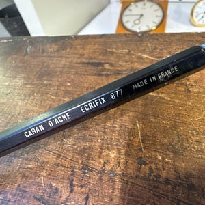 Könnte beinhalten: Ein schwarzer Caran d'Ache Ecrifix 877 Stift mit weißem Aufdruck CARAN D'ACHE ECRIFIX 877 MADE IN FRANCE. Der Stift hat einen sechseckigen Schaft und ein kleines blaues Bauteil ist an der Spitze des Schreibgeräts deutlich zu erkennen.
