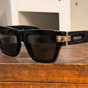 May include: Black sunglasses with dark lenses, featuring a thick, rectangular frame. Gold accents are visible on the temples, along with the brand name "Versace". The sunglasses are placed on a wooden surface.