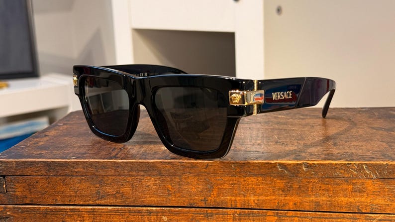 May include: Black sunglasses with dark lenses, featuring a thick, rectangular frame. Gold accents are visible on the temples, along with the brand name "Versace". The sunglasses are placed on a wooden surface.