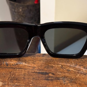 May include: Black sunglasses with a thick, rectangular frame and dark lenses. The temples are attached with small gold-colored accents. The sunglasses are resting on a wooden surface.