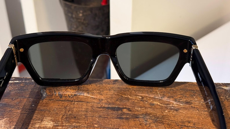 May include: Black sunglasses with a thick, rectangular frame and dark lenses. The temples are attached with small gold-colored accents. The sunglasses are resting on a wooden surface.