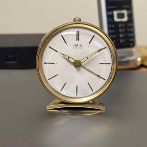 Könnte beinhalten: Ein goldfarbener, vintage Oris Wecker mit weißem Zifferblatt und schwarzen Stundenmarkierungen. Die Uhr hat goldene Zeiger und einen goldenen Sockel. Der Text "ORIS 7 JEWELS" ist auf dem Zifferblatt aufgedruckt. Die Uhr steht auf einer grauen Oberfläche.