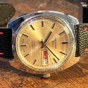 May include: A silver-toned wristwatch with a black leather strap and red stitching. The watch face is gold-coloured with black hands and markers. The watch face also has a date window and the text "DELVINA Automatic Incabloc Geneva" printed on it.