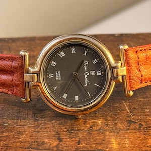 May include: A Pierre Cardin wristwatch with a gold-tone case and a dark dial featuring Roman numeral hour markers. The watch has a brown leather strap. The watch face reads "Swiss Made".