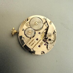 K&ouml;nnte beinhalten: Nahaufnahme eines Vintage-Uhrwerks, das komplizierte Zahnr&auml;der, Juwelen und eine Aufzugswelle zeigt. Der silberfarbene Mechanismus tr&auml;gt die Aufschrift "SWISS MADE" und "17 JEWELS". Es ist ein mechanisches Uhrwerk.