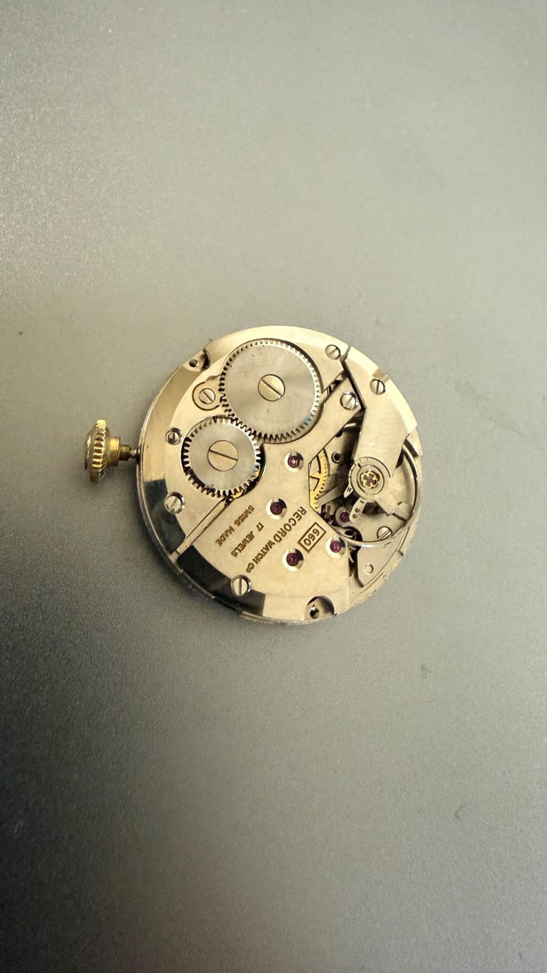 K&ouml;nnte beinhalten: Nahaufnahme eines Vintage-Uhrwerks, das komplizierte Zahnr&auml;der, Juwelen und eine Aufzugswelle zeigt. Der silberfarbene Mechanismus tr&auml;gt die Aufschrift "SWISS MADE" und "17 JEWELS". Es ist ein mechanisches Uhrwerk.