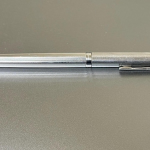 Può includere: Una penna a sfera argentata con finitura strutturata. La penna ha un design elegante e aerodinamico con una clip da taschino. La penna è su una superficie grigia.