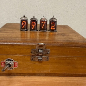 Op de afbeelding: Een rechthoekige houten doos met vier gloeiende oranje nixie-buizen op het bovenoppervlak die de cijfers 9, 9, 7 en 2 weergeven. De voorkant van de doos bevat een metalen grendel en een zilveren tuimelschakelaar met een plaatje met ON en OFF.