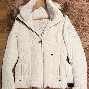 May include: White puffer jacket with a faux fur hood and a zip closure. The jacket has a quilted design and a black pocket on the left side. The text "POIVRE BLANC" is printed on a label on the front of the jacket.