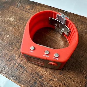 Op de afbeelding: Een rood polshorloge met een digitaal display en drie knoppen met de labels "SET", "RESET" en "SCAN". Het horloge heeft een zilveren metalen sluiting.