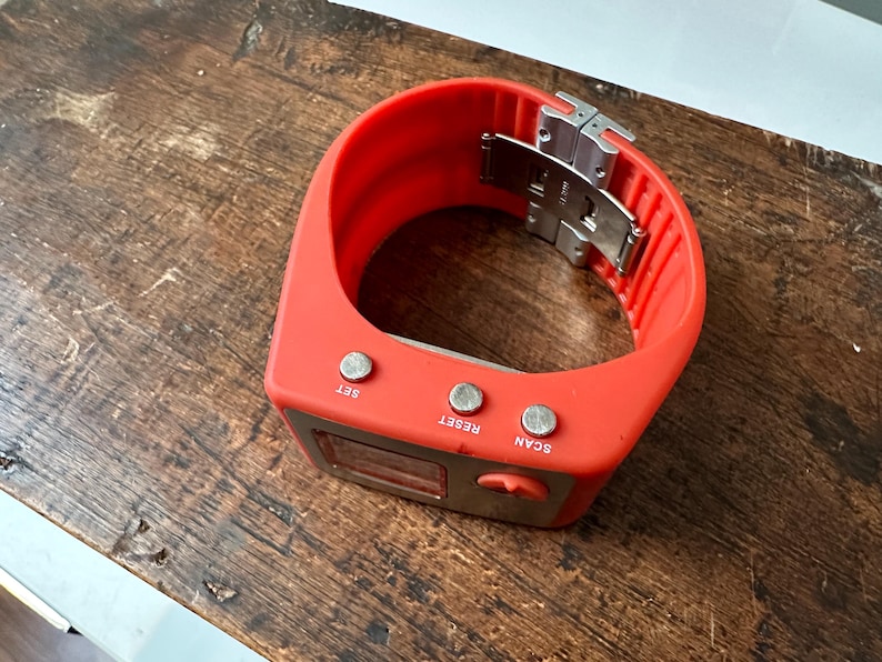 Op de afbeelding: Een rood polshorloge met een digitaal display en drie knoppen met de labels "SET", "RESET" en "SCAN". Het horloge heeft een zilveren metalen sluiting.