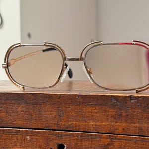 May include: Gold-rimmed sunglasses with brown-tinted lenses. The glasses have a unique, geometric shape with a double-bar design above the lenses. The temples are thin and black, and the glasses are resting on a wooden surface.
