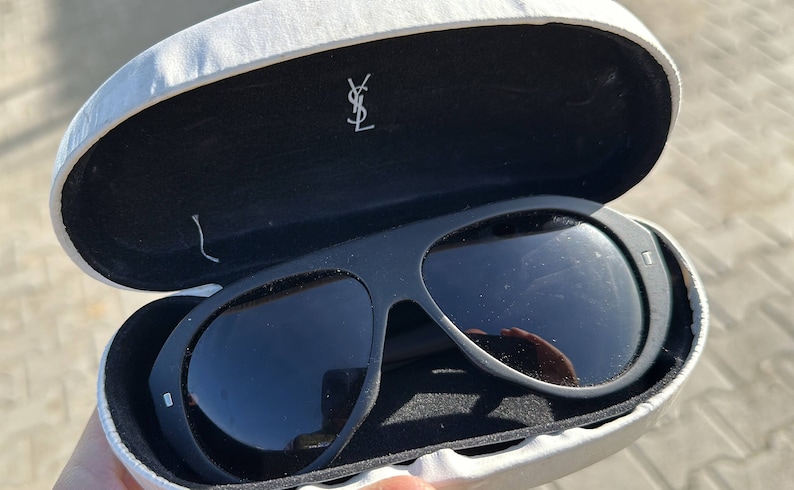 Pode incluir: &Oacute;culos de sol pretos com um logotipo YSL na caixa. Os &oacute;culos de sol est&atilde;o em uma caixa r&iacute;gida branca.
