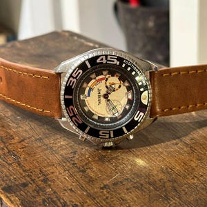 May include: A silver Jim Beam watch with a black face and brown leather band. The watch has a chronograph and a date window. The watch face has the words "Jim Beam" and "Additional Time" printed on it.