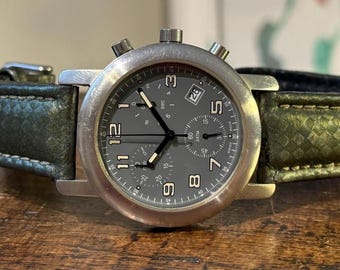 May include: A silver-toned wristwatch with a dark gray face and a green leather band. The watch features a date display and multiple sub-dials. The band is textured and stitched. The watch is displayed on a wooden surface.