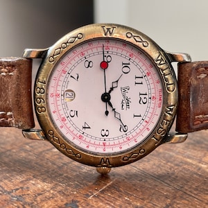 Pode incluir: Um relógio de pulso dourado com um mostrador branco e ponteiros pretos. O relógio tem uma pulseira de couro marrom e uma escala de tacômetro ao redor da borda do mostrador. O mostrador do relógio diz "Wild West" e "Telemeter".