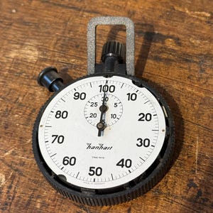 May include: A vintage black and white stopwatch with a metal handle. The watch face displays numbers from 0 to 100, with the brand name "Hanhart" visible. The stopwatch is likely used for timing events or measuring intervals.