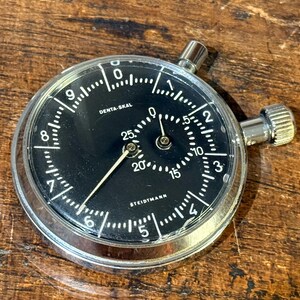 Könnte beinhalten: Eine Vintage Denta-Skal Stoppuhr mit schwarzem Zifferblatt und weißen Markierungen. Die Stoppuhr hat ein silbernes Metallgehäuse und zwei Aufzugsknöpfe. Das Zifferblatt zeigt den Markennamen Steidmann.