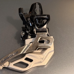 May include: A bicycle front derailleur with a silver and black finish. The component features the text "SRAM XO" and is designed for mountain bikes. The derailleur is made of metal and has a complex mechanical design.