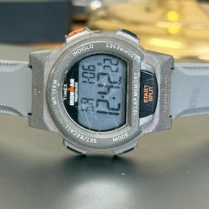 K&ouml;nnte beinhalten: Eine graue Timex Ironman Triathlon Digitaluhr mit grauem Armband. Das Zifferblatt zeigt die Uhrzeit 12:42:22 an. Die Uhr hat mehrere Kn&ouml;pfe und den Text "INDIGLO" und "WR 100M".