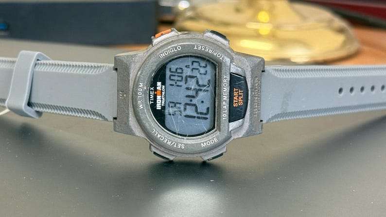 K&ouml;nnte beinhalten: Eine graue Timex Ironman Triathlon Digitaluhr mit grauem Armband. Das Zifferblatt zeigt die Uhrzeit 12:42:22 an. Die Uhr hat mehrere Kn&ouml;pfe und den Text "INDIGLO" und "WR 100M".