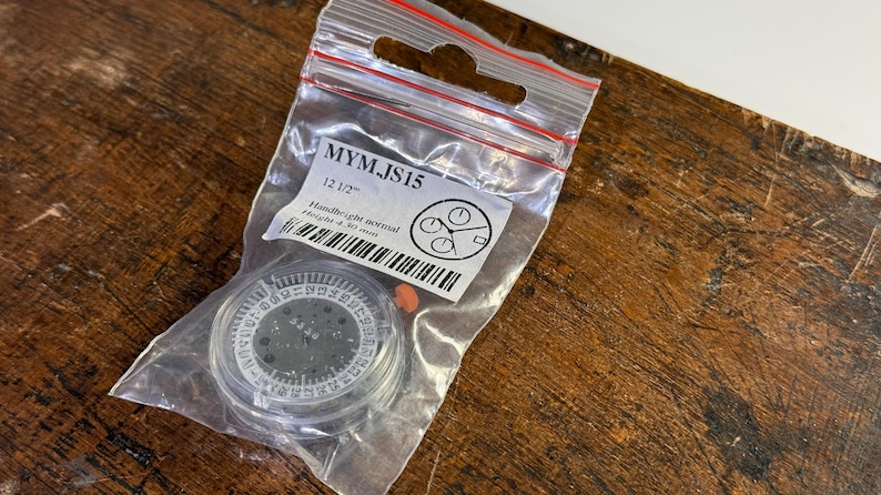 May include: A clear plastic bag containing a watch face with black numbers and a silver ring. The bag also contains a small orange sticker and a white label with the text "MYM_JS15 12 1/2" Hand-light control 1/10 of a sec.