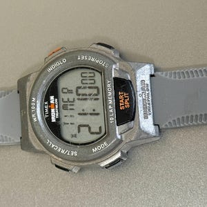 K&ouml;nnte beinhalten: Eine graue Timex Ironman Digitaluhr mit grauem Armband. Das Zifferblatt zeigt die Zeit 2:10:00 an. Funktionen: Stoppuhr, Rundenspeicher und Indiglo-Hintergrundbeleuchtung. Die Uhr ist f&uuml;r das Triathlon-Training konzipiert.