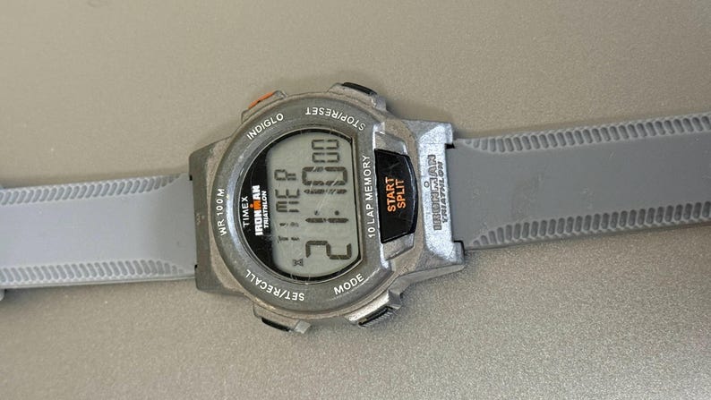 K&ouml;nnte beinhalten: Eine graue Timex Ironman Digitaluhr mit grauem Armband. Das Zifferblatt zeigt die Zeit 2:10:00 an. Funktionen: Stoppuhr, Rundenspeicher und Indiglo-Hintergrundbeleuchtung. Die Uhr ist f&uuml;r das Triathlon-Training konzipiert.