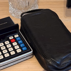 May include: A black electronic calculator with a silver trim and a black leather case. The calculator has a display screen, number keys, and function keys. The case has a zipper closure.