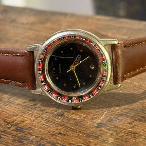 Pode incluir: Um relógio de pulso vintage com um mostrador preto com um design de roleta. O relógio tem uma caixa prateada, ponteiros dourados e uma pulseira de couro castanho. O mostrador exibe números e a palavra "QUARTZ".