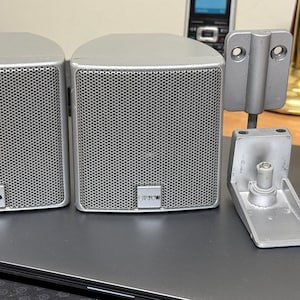 May include: Two silver Canton speakers with a mesh front, and two sets of silver mounting brackets. The speakers have a rectangular shape with rounded tops. The Canton logo is visible on the front of each speaker.