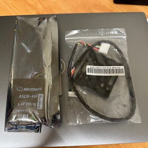 May include: Two sealed plastic bags containing electronic components. One bag holds a rectangular component labeled "Microsemi ASCM-35F 5.4V 35F". The other bag contains a black bracket with wires and a label reading "ASM-00631-02-A-R REV A3".