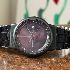 May include: A black Junghans Solar 1 wristwatch with a dark purple face and a black band. The watch has a date display and red second hand. The watch is on a wooden surface.