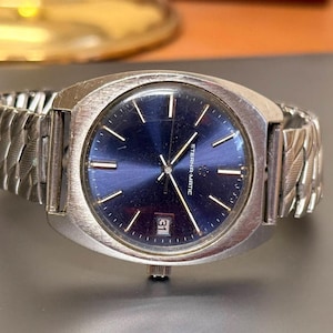 Puede incluir: Reloj de pulsera Eterna-Matic plateado con esfera azul y correa plateada. La esfera del reloj tiene marcadores y agujas plateadas, con una visualización de la fecha a las 3 en punto. La correa es de diseño texturizado y enlazado.