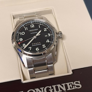May include: A stainless steel Longines automatic watch with a black dial, white hands, and Arabic numeral hour markers. The watch is presented in a cream-colored box with the Longines logo visible.