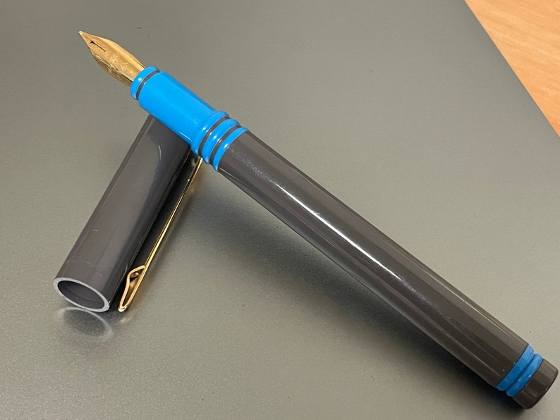 May include: A gray and blue fountain pen with a gold nib and clip. The pen is closed and the cap is off.