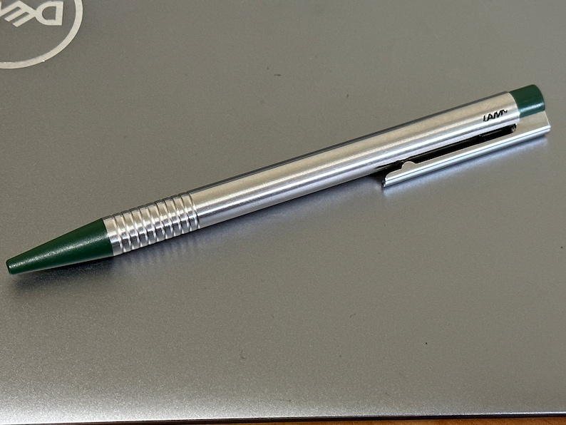 Puede incluir: Un bol&iacute;grafo plateado con un capuch&oacute;n verde y una punta verde. El bol&iacute;grafo tiene un cuerpo acanalado y un clip en el lateral. El bol&iacute;grafo est&aacute; hecho por LAMY.