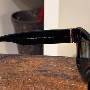 May include: Black sunglasses with a rectangular frame. The temple arm features the text "MOD. 4464 GB1/87 55020 145 3N". The sunglasses are resting on a wooden surface.