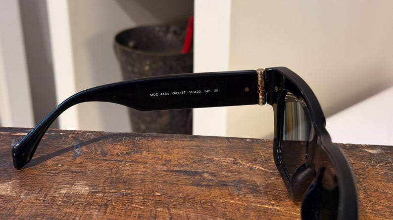 May include: Black sunglasses with a rectangular frame. The temple arm features the text "MOD. 4464 GB1/87 55020 145 3N". The sunglasses are resting on a wooden surface.