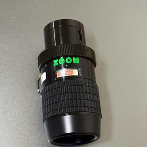 Puede incluir: Un objetivo zoom negro con texto verde "ZOOM" y "X 22X". El objetivo tiene un agarre estriado y un anillo de ajuste plateado. El objetivo est&aacute; dise&ntilde;ado para fotograf&iacute;a.