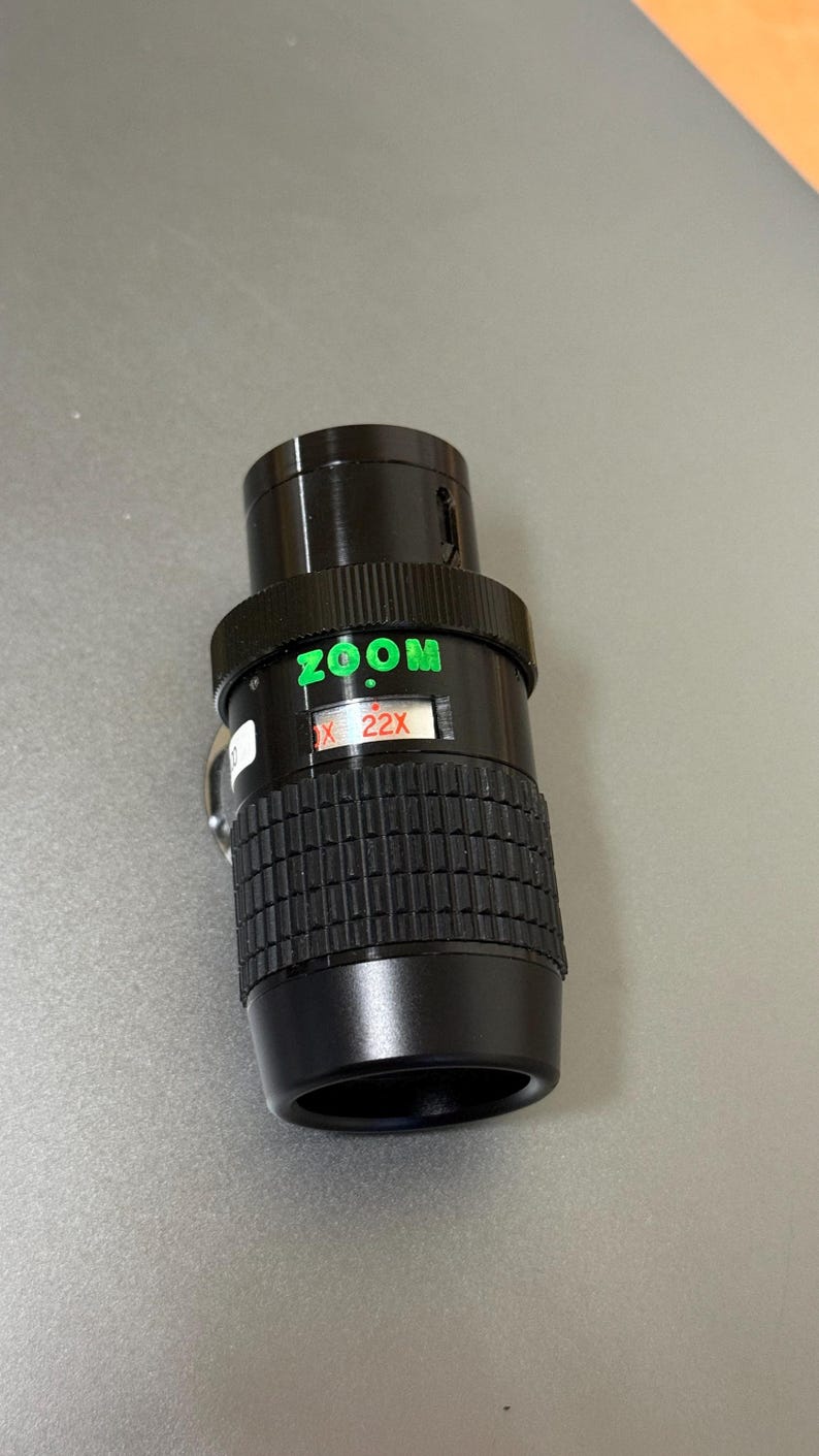 Puede incluir: Un objetivo zoom negro con texto verde "ZOOM" y "X 22X". El objetivo tiene un agarre estriado y un anillo de ajuste plateado. El objetivo est&aacute; dise&ntilde;ado para fotograf&iacute;a.