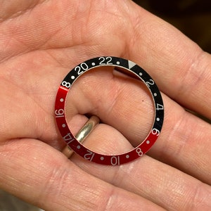 May include: A black and red watch bezel with white numbers and a white triangle at the 12 o'clock position.