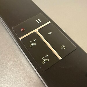 Pode incluir: Um painel de controle preto com vários botões. Os botões incluem ligar/desligar, aumentar e diminuir a velocidade da ventoinha, setas para cima e para baixo, um botão "int" e um temporizador. Os botões têm ícones e texto brancos.