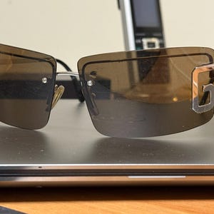 May include: Rimless sunglasses with brown tinted lenses. The sunglasses have a silver frame and a decorative silver "G" on the temples. The sunglasses are resting on a dark gray surface.