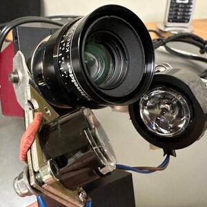Op de afbeelding: Een close-up van een zwarte camera lens met een zilveren ring en markeringen die "Schneider - Kreuznach 1:1.4 28 mm" lezen. De lens is bevestigd aan een zilveren en zwarte metalen beugel met een rode draad eraan bevestigd. Rechts van de camera lens is er een zwarte en zilveren lichtarmatuur met een heldere lens.