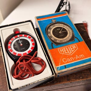 以下が含まれることがあります： オリジナルの箱に入った、ヴィンテージのHeuer Can-Amストップウォッチ。ストップウォッチは、黒い文字盤に赤と白のマーキング、赤いコードが付いています。箱は青とオレンジで、Heuerのロゴと「Can-Am」の文字が入っています。