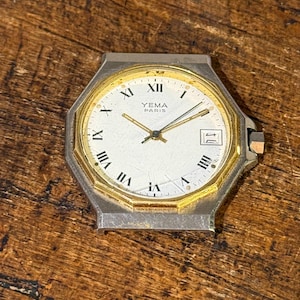 May include: Vintage Yema Paris watch with a silver-tone case and a gold-tone bezel. The watch face is white with black Roman numerals and gold hands. The watch has a date window at the 3 o'clock position. The watch is displayed on a wooden surface.