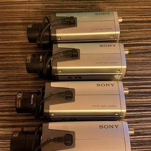 May include: Four silver Sony colour video cameras are arranged in a row. Each camera has a black lens and a black cable. The text "SONY" and "COLOR VIDEO CAMERA" are printed on the front of each camera.