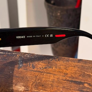 May include: Black Versace sunglasses with gold accents. The text "VERSACE MADE IN ITALY" is printed on the arm. A red stripe is visible on the arm. The sunglasses are resting on a wooden surface.